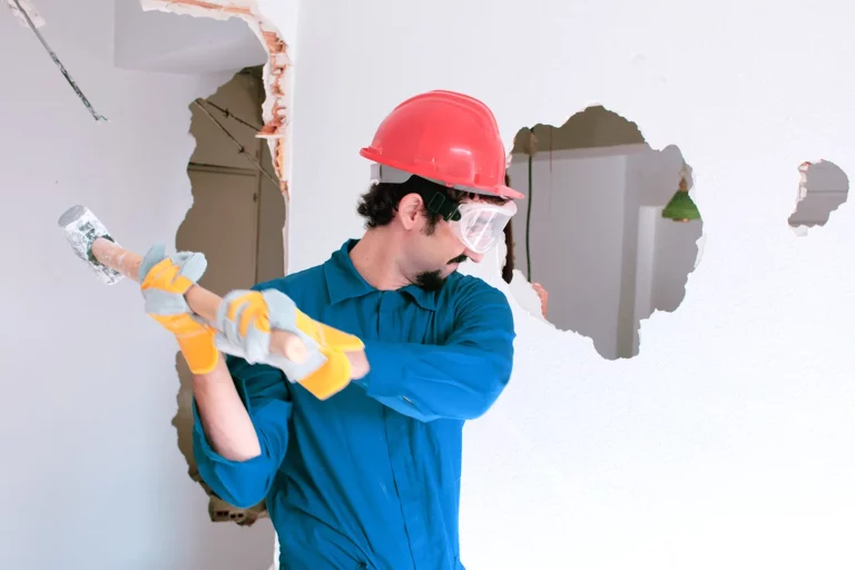 A worker in a red helmet and blue overalls dismantles a wall